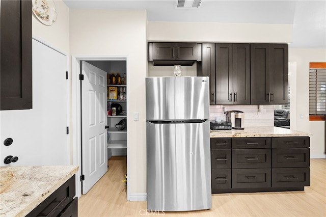 kitchen with light stone countertops, backsplash, dark brown cabinetry, light hardwood / wood-style floors, and stainless steel refrigerator