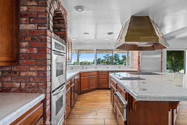 kitchen with sink, light wood-type flooring, tile counters, island exhaust hood, and stainless steel appliances