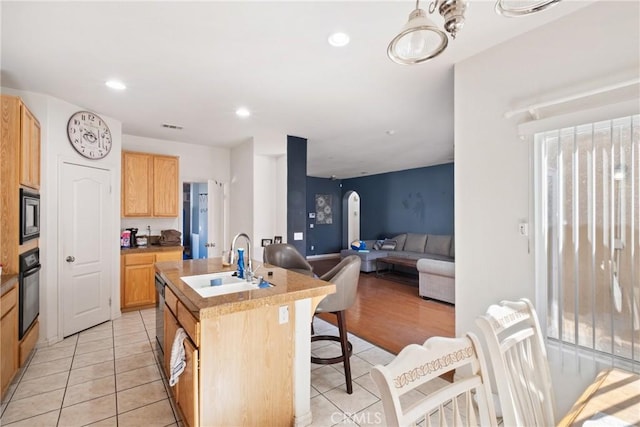 kitchen featuring stainless steel microwave, sink, black oven, a kitchen island with sink, and light tile patterned flooring