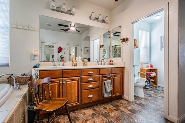 bathroom with ceiling fan, tiled tub, toilet, and vanity