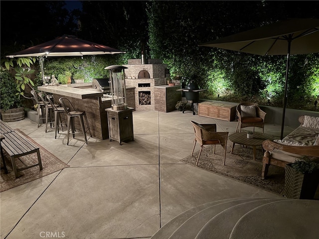 patio at twilight with an outdoor kitchen, a grill, exterior bar, and an outdoor stone fireplace