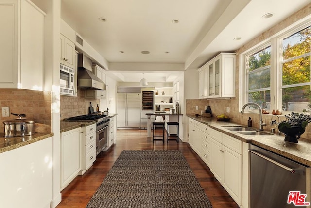 kitchen featuring dark wood-type flooring, wall chimney range hood, sink, built in appliances, and white cabinetry