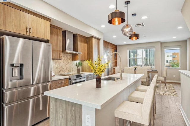 kitchen with wall chimney exhaust hood, sink, a center island with sink, pendant lighting, and stainless steel appliances