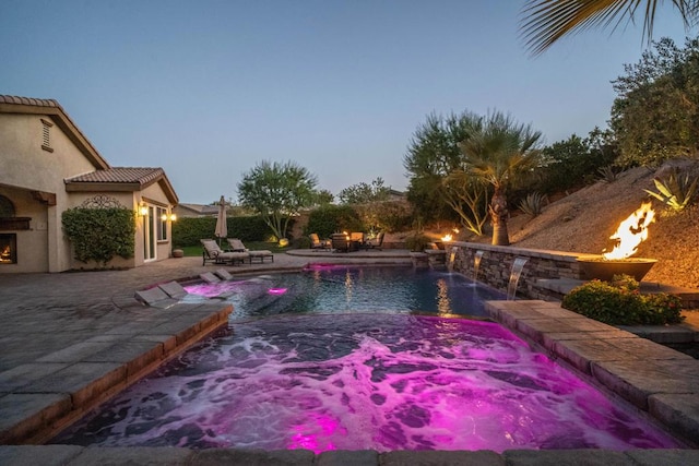 pool at dusk with exterior fireplace, pool water feature, and a patio