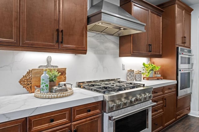 kitchen with appliances with stainless steel finishes, backsplash, dark hardwood / wood-style flooring, light stone countertops, and wall chimney range hood