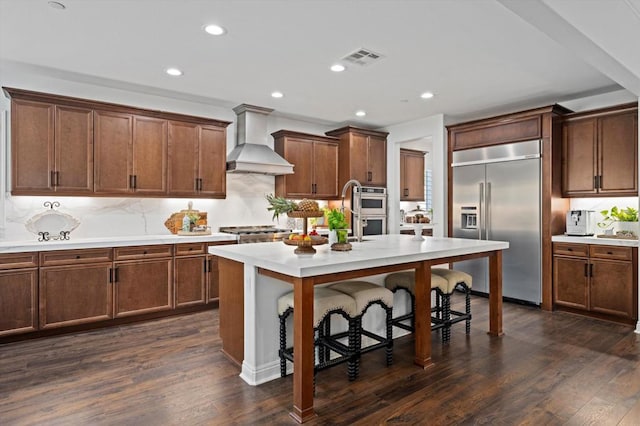 kitchen with tasteful backsplash, dark hardwood / wood-style floors, a kitchen island with sink, appliances with stainless steel finishes, and custom range hood