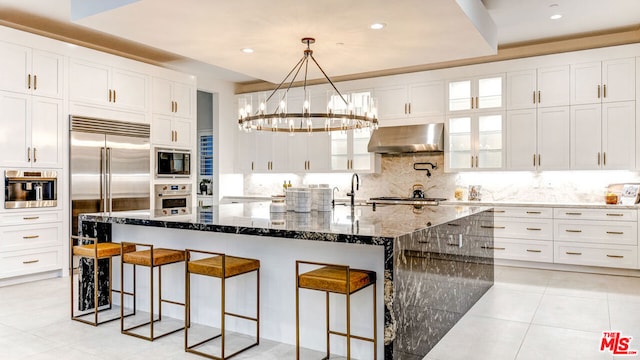 kitchen with extractor fan, a center island with sink, dark stone countertops, built in appliances, and a breakfast bar