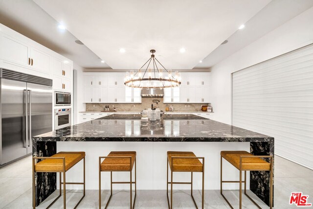 kitchen with a breakfast bar, a large island with sink, dark stone counters, and built in appliances