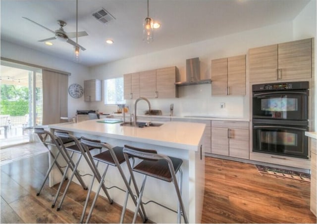 kitchen with sink, wall chimney exhaust hood, black double oven, decorative light fixtures, and a kitchen island with sink