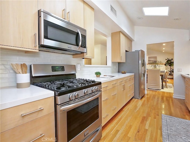 kitchen featuring light brown cabinets, stainless steel appliances, backsplash, vaulted ceiling, and light wood-type flooring