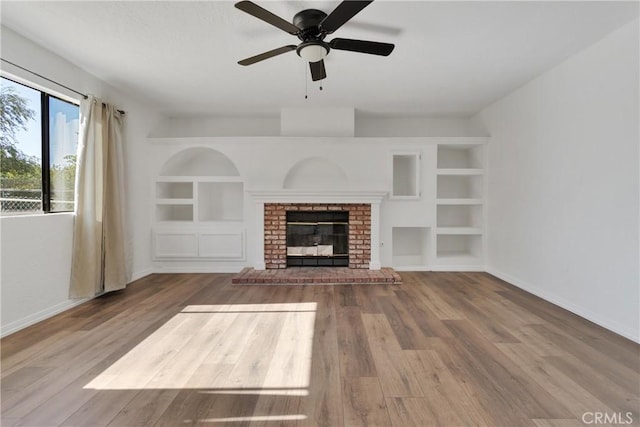 unfurnished living room featuring built in features, wood-type flooring, a brick fireplace, and ceiling fan