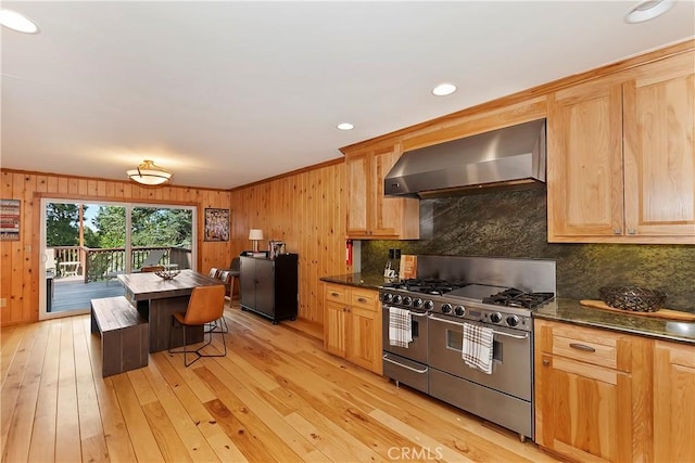 kitchen featuring light brown cabinets, range with two ovens, light wood-type flooring, dark stone counters, and wall chimney exhaust hood