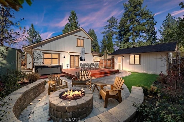 back house at dusk featuring a wooden deck, a patio area, a hot tub, and a fire pit