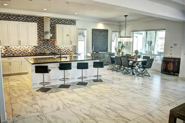 kitchen with white cabinetry, hanging light fixtures, an island with sink, a breakfast bar area, and wall chimney exhaust hood