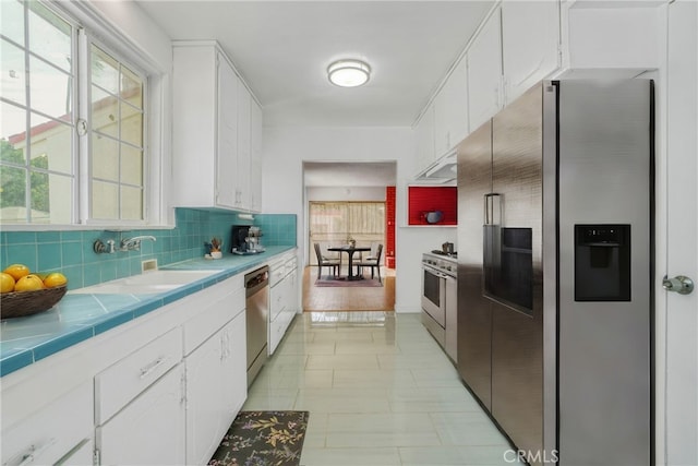 kitchen with appliances with stainless steel finishes, sink, tile countertops, white cabinets, and light hardwood / wood-style floors