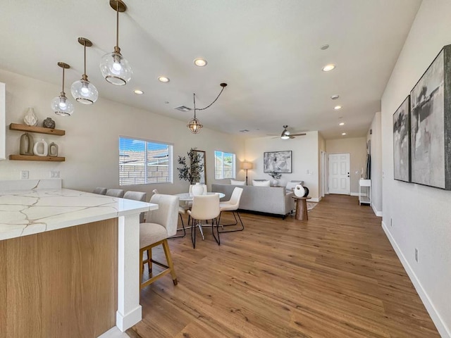 kitchen with ceiling fan, light stone counters, wood-type flooring, decorative light fixtures, and a breakfast bar area