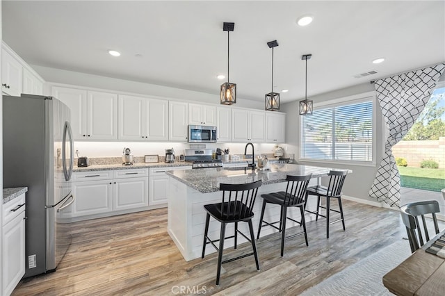 kitchen featuring white cabinetry, an island with sink, appliances with stainless steel finishes, light wood-type flooring, and hanging light fixtures