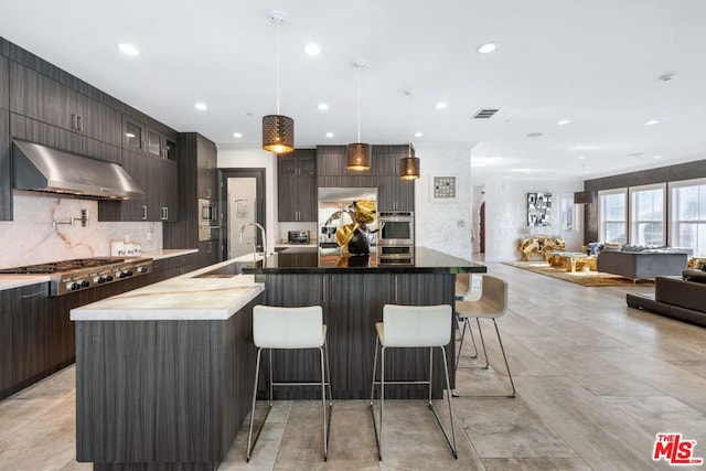 kitchen featuring wall chimney exhaust hood, a kitchen island with sink, sink, built in appliances, and decorative light fixtures