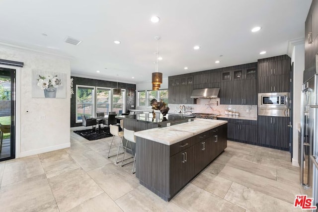 kitchen with stainless steel appliances, hanging light fixtures, tasteful backsplash, a kitchen island with sink, and exhaust hood