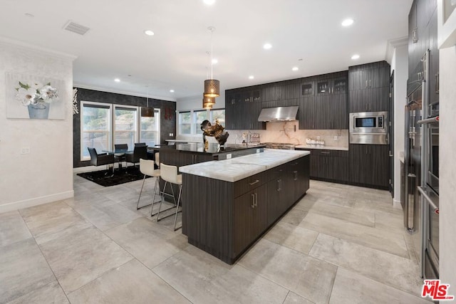 kitchen featuring hanging light fixtures, wall chimney exhaust hood, decorative backsplash, appliances with stainless steel finishes, and a kitchen island