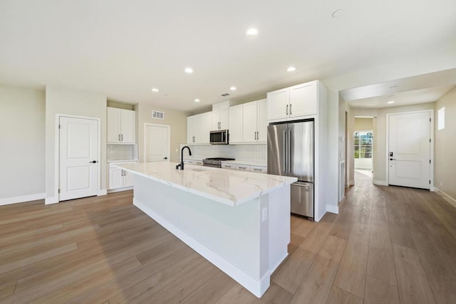 kitchen featuring light stone countertops, appliances with stainless steel finishes, a center island with sink, light hardwood / wood-style floors, and white cabinetry