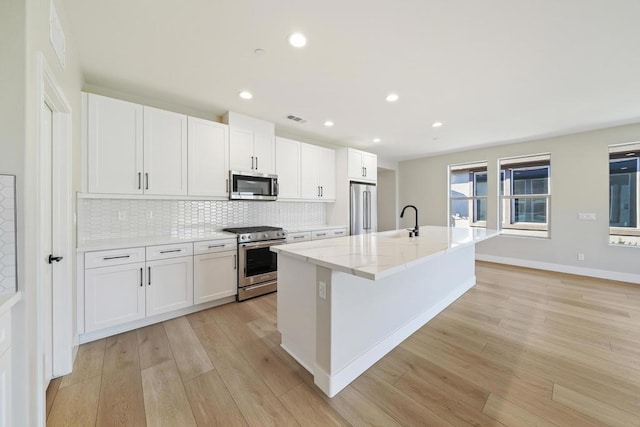 kitchen with light stone countertops, light wood-type flooring, an island with sink, appliances with stainless steel finishes, and white cabinetry