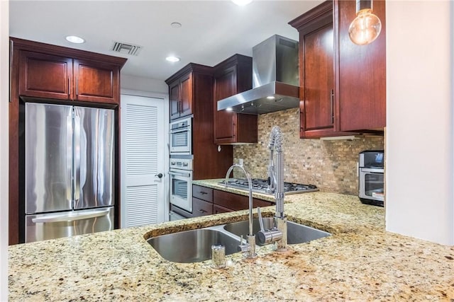 kitchen with light stone countertops, appliances with stainless steel finishes, backsplash, wall chimney range hood, and decorative light fixtures