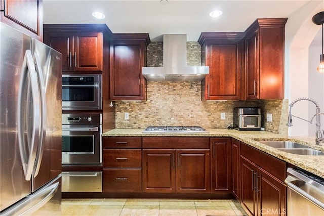 kitchen with light stone countertops, appliances with stainless steel finishes, wall chimney exhaust hood, sink, and decorative light fixtures