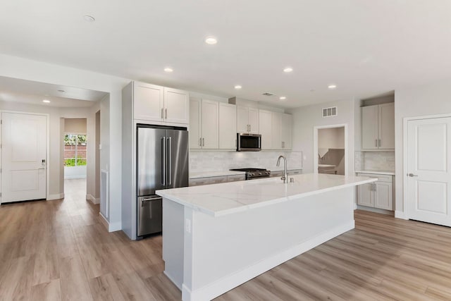 kitchen with decorative backsplash, light wood-type flooring, stainless steel appliances, a center island with sink, and white cabinetry