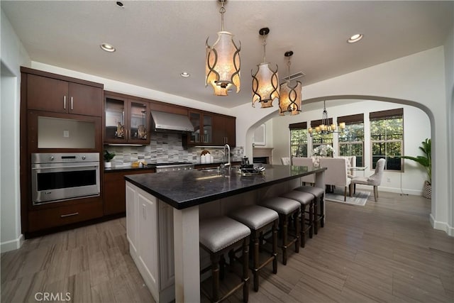 kitchen featuring stainless steel appliances, decorative light fixtures, a kitchen island with sink, dark brown cabinets, and exhaust hood