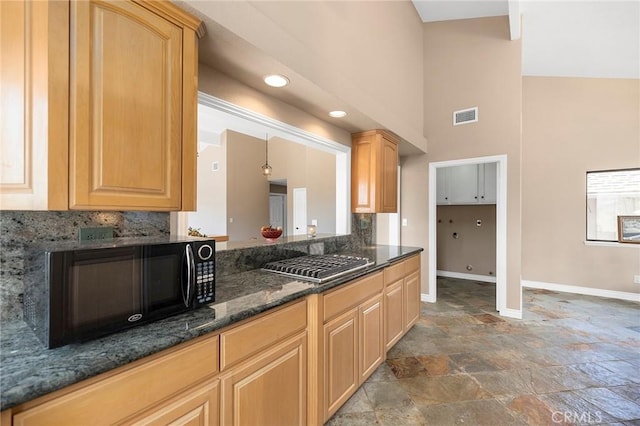 kitchen with light brown cabinetry, decorative light fixtures, dark stone countertops, decorative backsplash, and a high ceiling