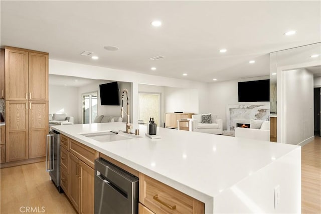 kitchen with a large island with sink, stainless steel dishwasher, sink, a premium fireplace, and light hardwood / wood-style flooring