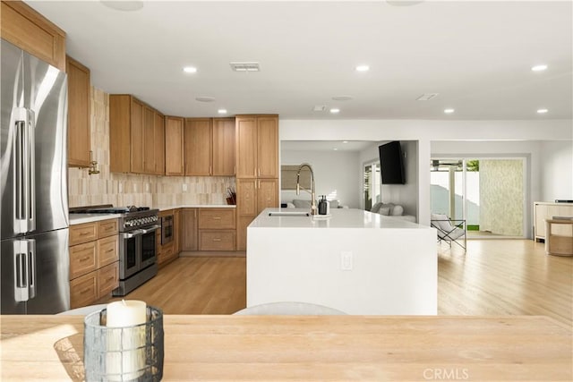 kitchen with an island with sink, decorative backsplash, high end appliances, light hardwood / wood-style flooring, and sink