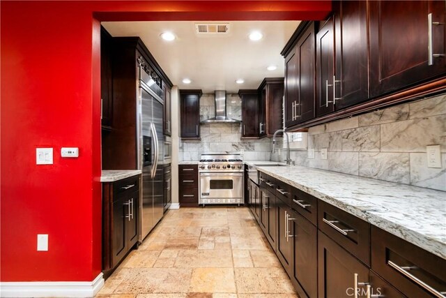 kitchen with light stone countertops, high end range, wall chimney exhaust hood, tasteful backsplash, and sink