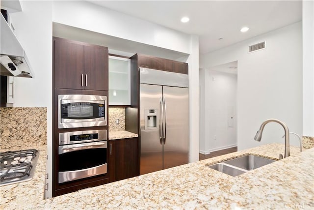 kitchen featuring backsplash, ventilation hood, sink, built in appliances, and light stone counters