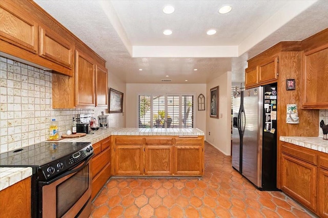 kitchen with tile countertops, kitchen peninsula, stainless steel appliances, backsplash, and a tray ceiling