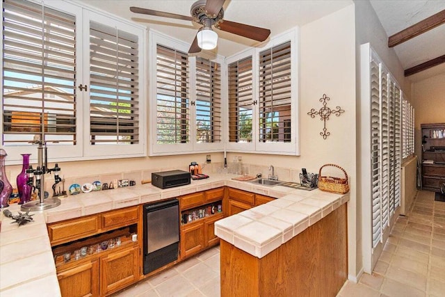 kitchen featuring ceiling fan, sink, beamed ceiling, tile counters, and light tile patterned floors