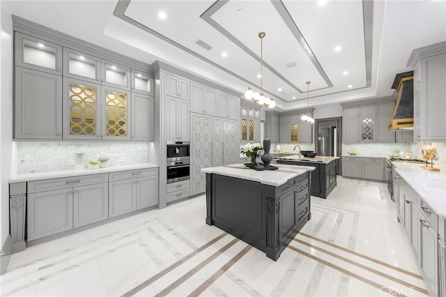 kitchen featuring backsplash, gray cabinets, a kitchen island with sink, and a tray ceiling