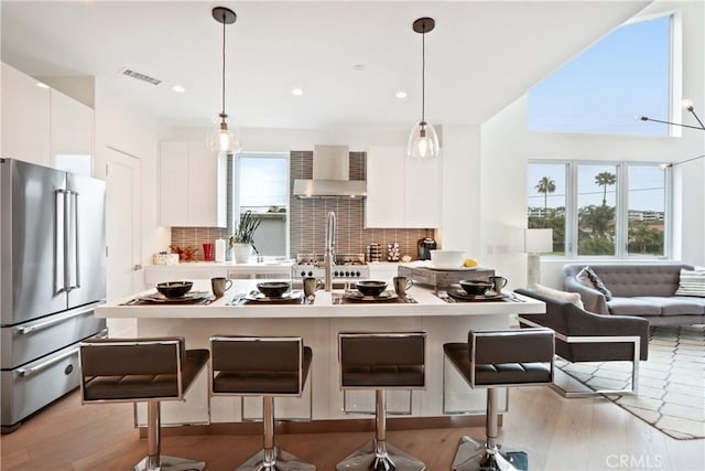 kitchen featuring decorative backsplash, stainless steel fridge, wall chimney exhaust hood, decorative light fixtures, and white cabinetry