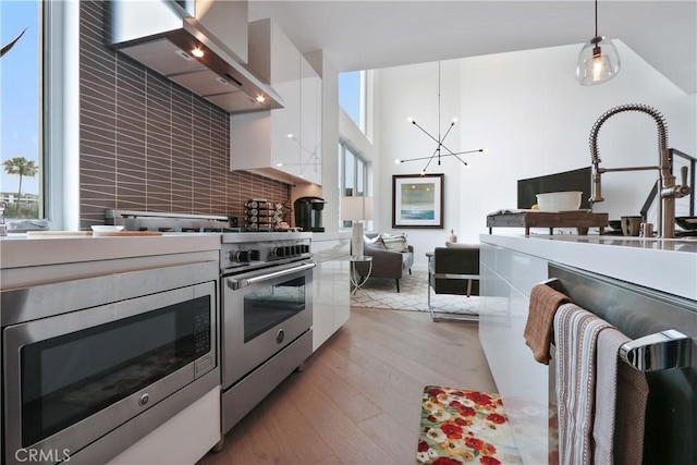 kitchen featuring light wood-type flooring, stainless steel appliances, wall chimney range hood, decorative light fixtures, and white cabinets