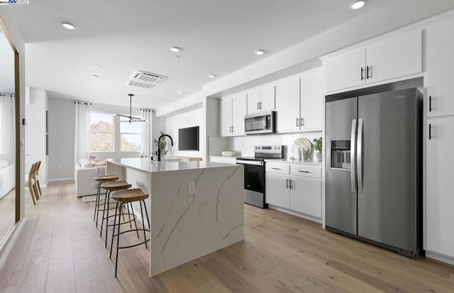 kitchen with hanging light fixtures, stainless steel appliances, an island with sink, white cabinets, and light wood-type flooring