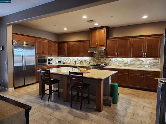 kitchen with a breakfast bar area, built in appliances, decorative backsplash, and an island with sink