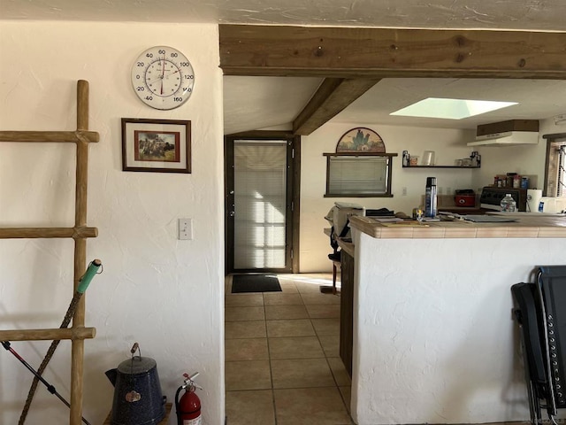 kitchen featuring tile countertops, stove, lofted ceiling with skylight, exhaust hood, and light tile patterned floors