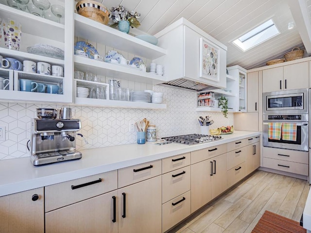 kitchen featuring vaulted ceiling with skylight, decorative backsplash, custom exhaust hood, stainless steel appliances, and light hardwood / wood-style flooring