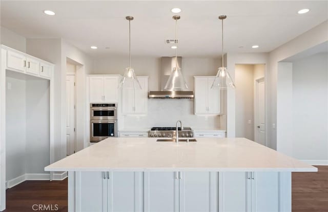 kitchen with sink, wall chimney range hood, white cabinets, hanging light fixtures, and an island with sink