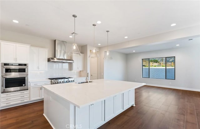 kitchen with wall chimney exhaust hood, stainless steel appliances, a kitchen island with sink, decorative light fixtures, and white cabinets