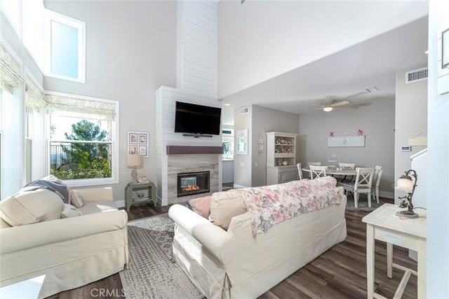 living room with ceiling fan, a large fireplace, a towering ceiling, and dark wood-type flooring