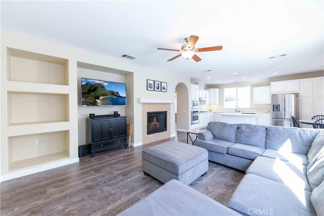 living room featuring built in shelves, ceiling fan, dark wood-type flooring, sink, and a tiled fireplace