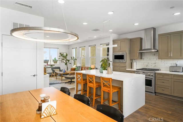 kitchen featuring wall chimney range hood, a kitchen breakfast bar, dark hardwood / wood-style floors, a kitchen island with sink, and appliances with stainless steel finishes