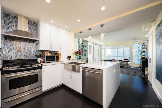 kitchen with dark wood-type flooring, wall chimney range hood, kitchen peninsula, pendant lighting, and appliances with stainless steel finishes